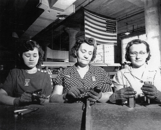 Três mulheres inspecionando cartuchos de munição, ao fundo uma bandeira dos Estados Unidos pendurada no teto.