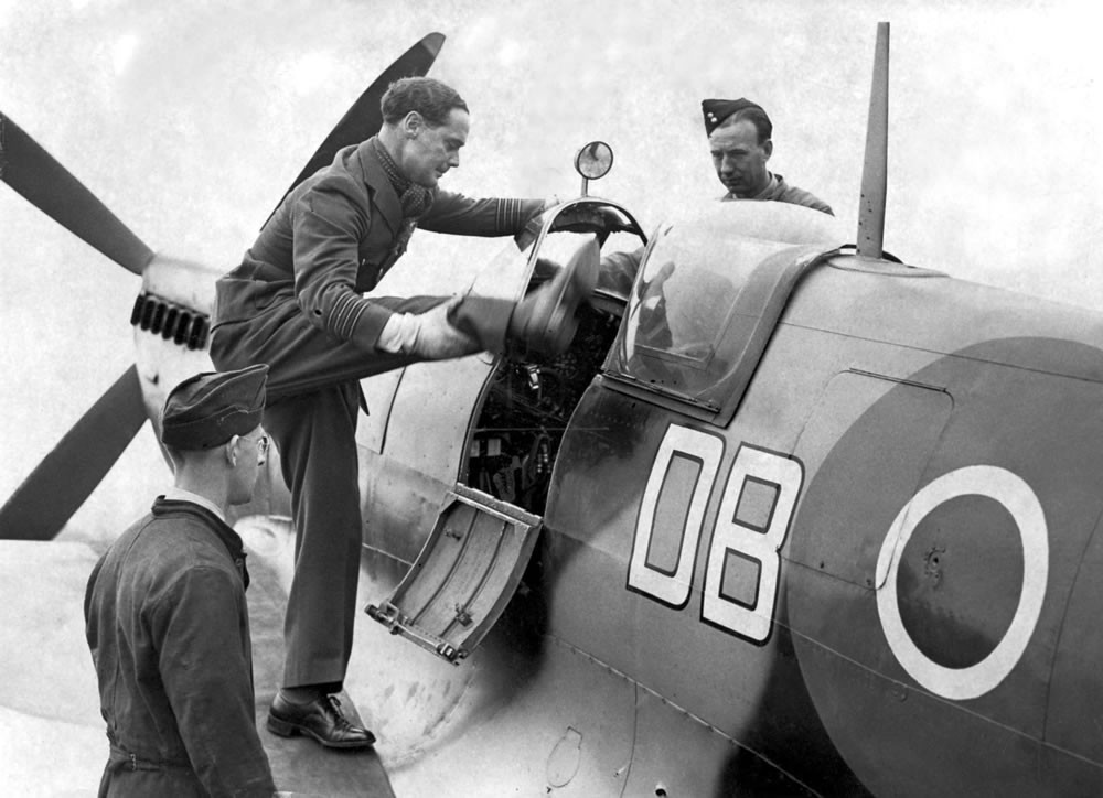 Douglas Bader em pé na asa de seu caça, segurando no vidro do cockpit, equilibrado em uma perna mecânica e levantando a outra para entrar no avião. 